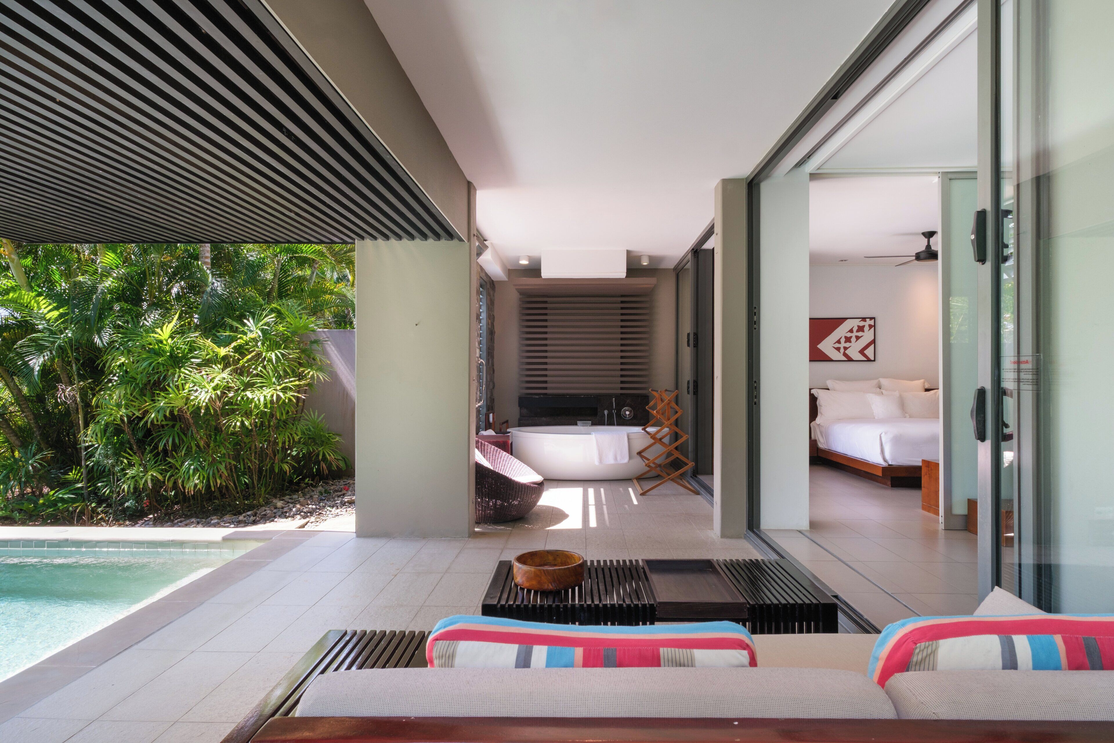 Une chambre d’hôtel moderne dotée d’une grande porte vitrée donnant sur un balcon avec piscine, une salle de bain privée et une chambre à coucher avec un lit et un ventilateur de plafond.