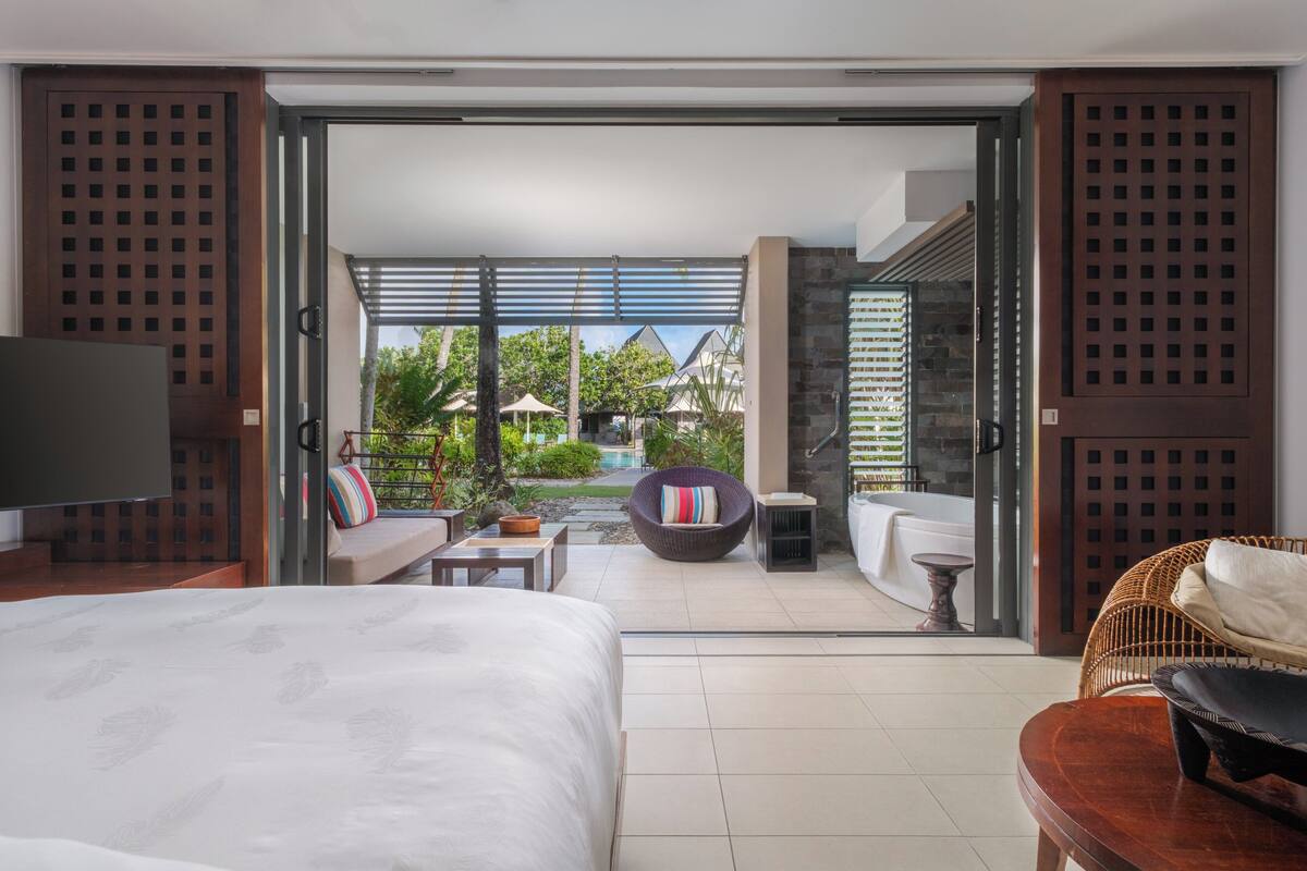 Ein modernes Schlafzimmer mit einem großen Bett, einem Fernseher und Blick auf einen Pool sowie eine Sitzgelegenheit im Freien.