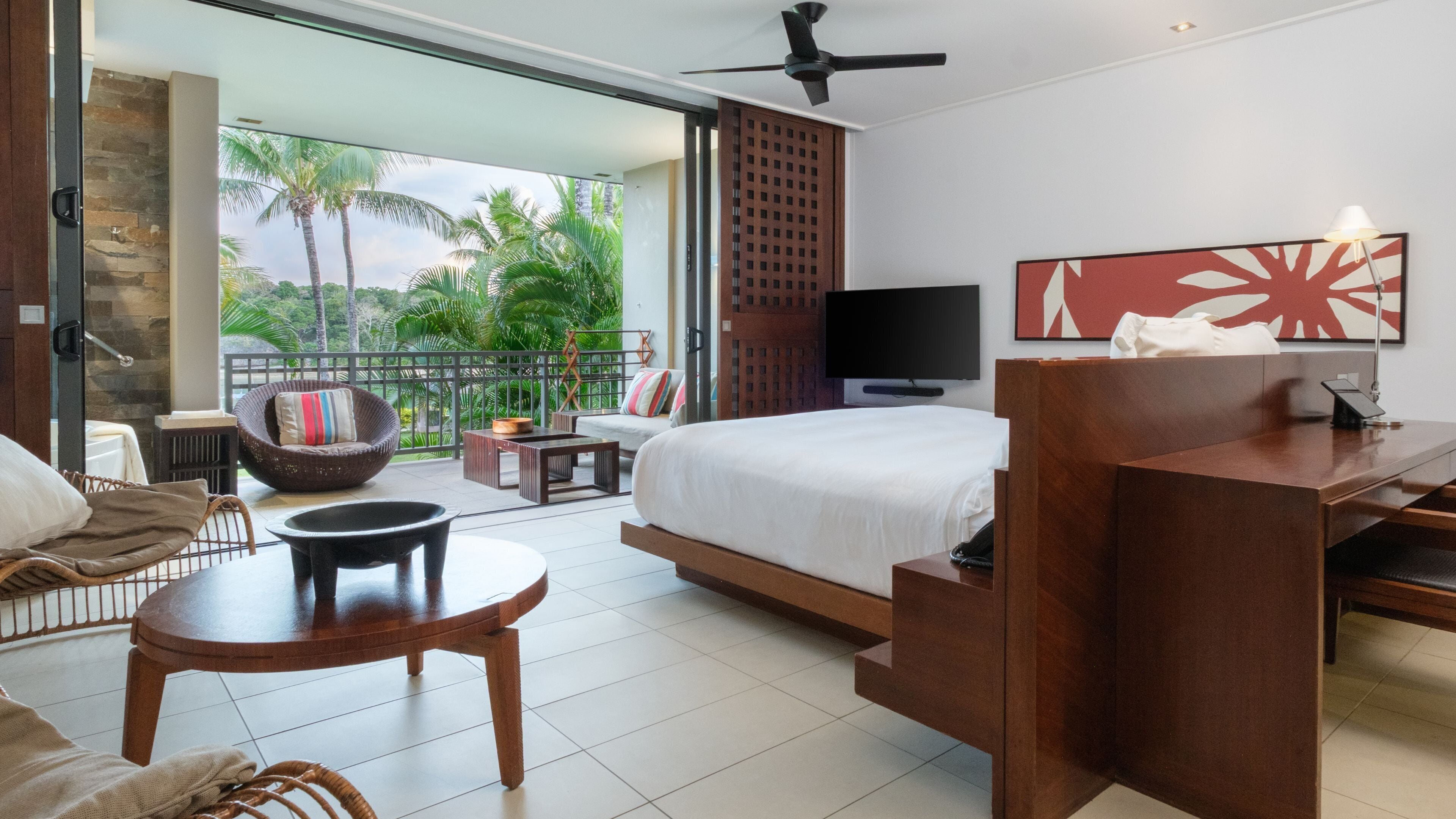 A modern hotel room with a bed, a desk, a chair, a small table, a sofa, and a balcony with a view of palm trees.