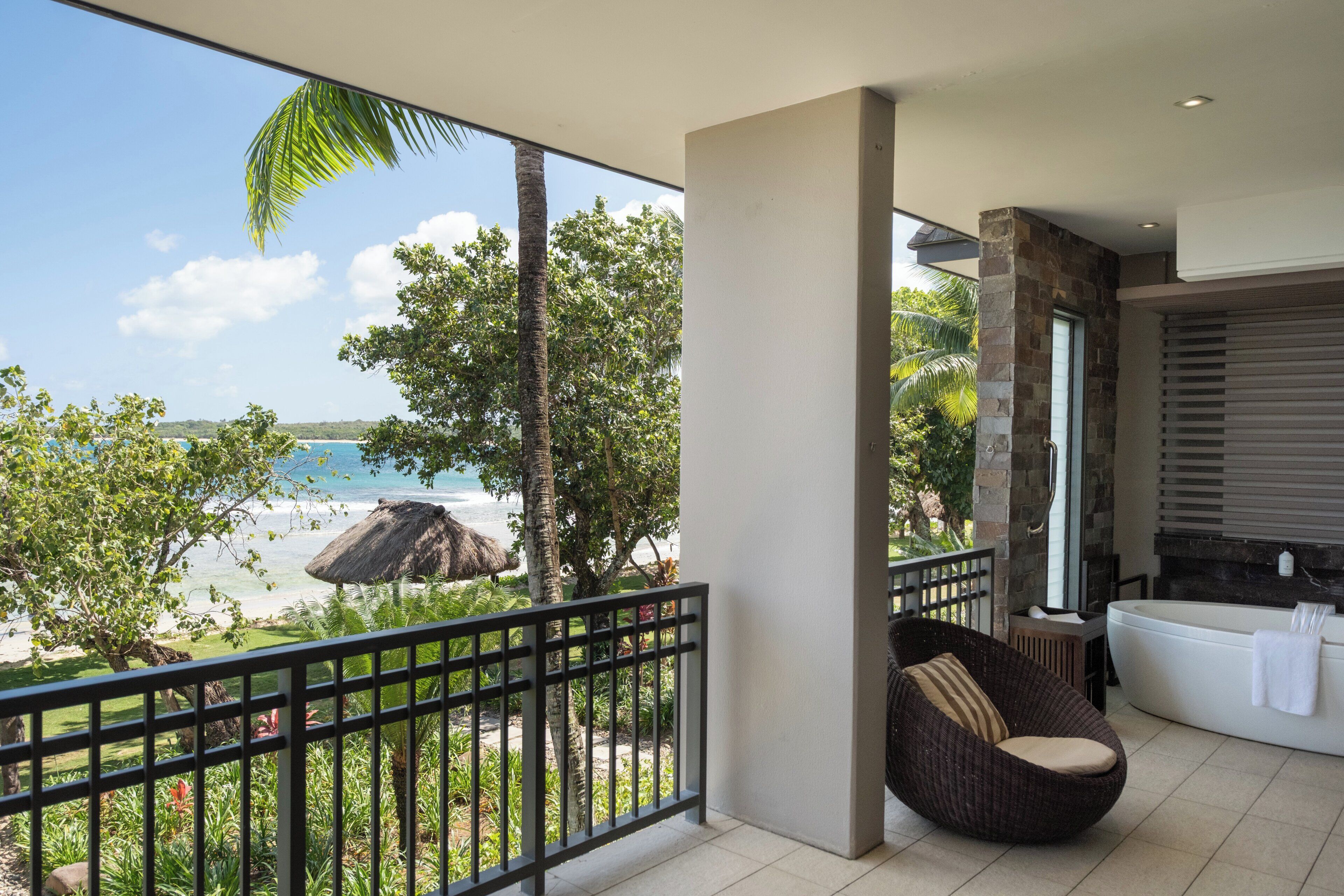 A balcony with a view of the beach and ocean, a wicker chair, and a bathtub.