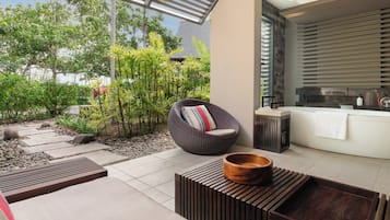 Un espacio exterior moderno con una zona de estar, una silla acolchada, una mesa de madera y una bañera con una toalla.