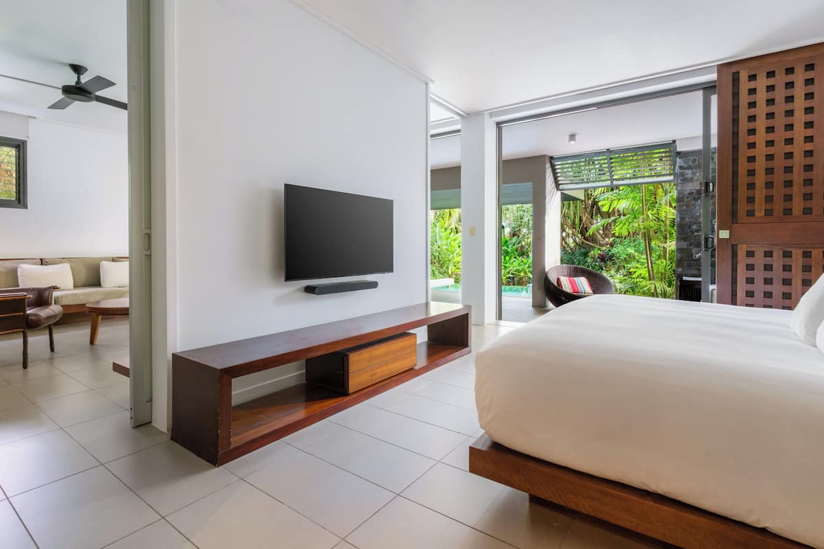 Ein modernes Schlafzimmer mit einem großen Bett, einem flachbildfernseher an einer Holzkonsole und einer Glastür, die auf einen Balkon mit üppiger Bepflanzung führt.