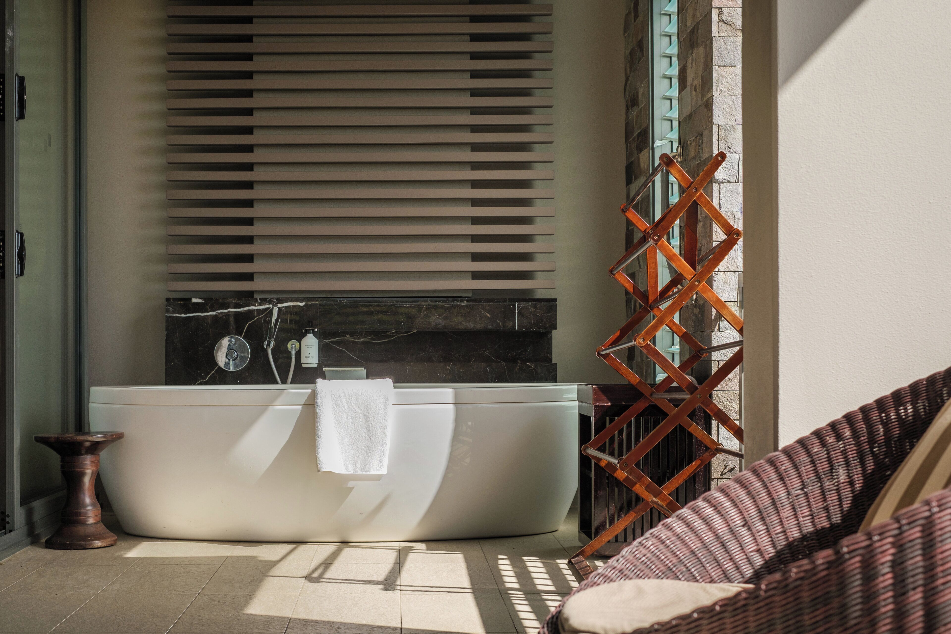 A modern bathroom with a freestanding bathtub, a wooden stool, and a wicker chair.