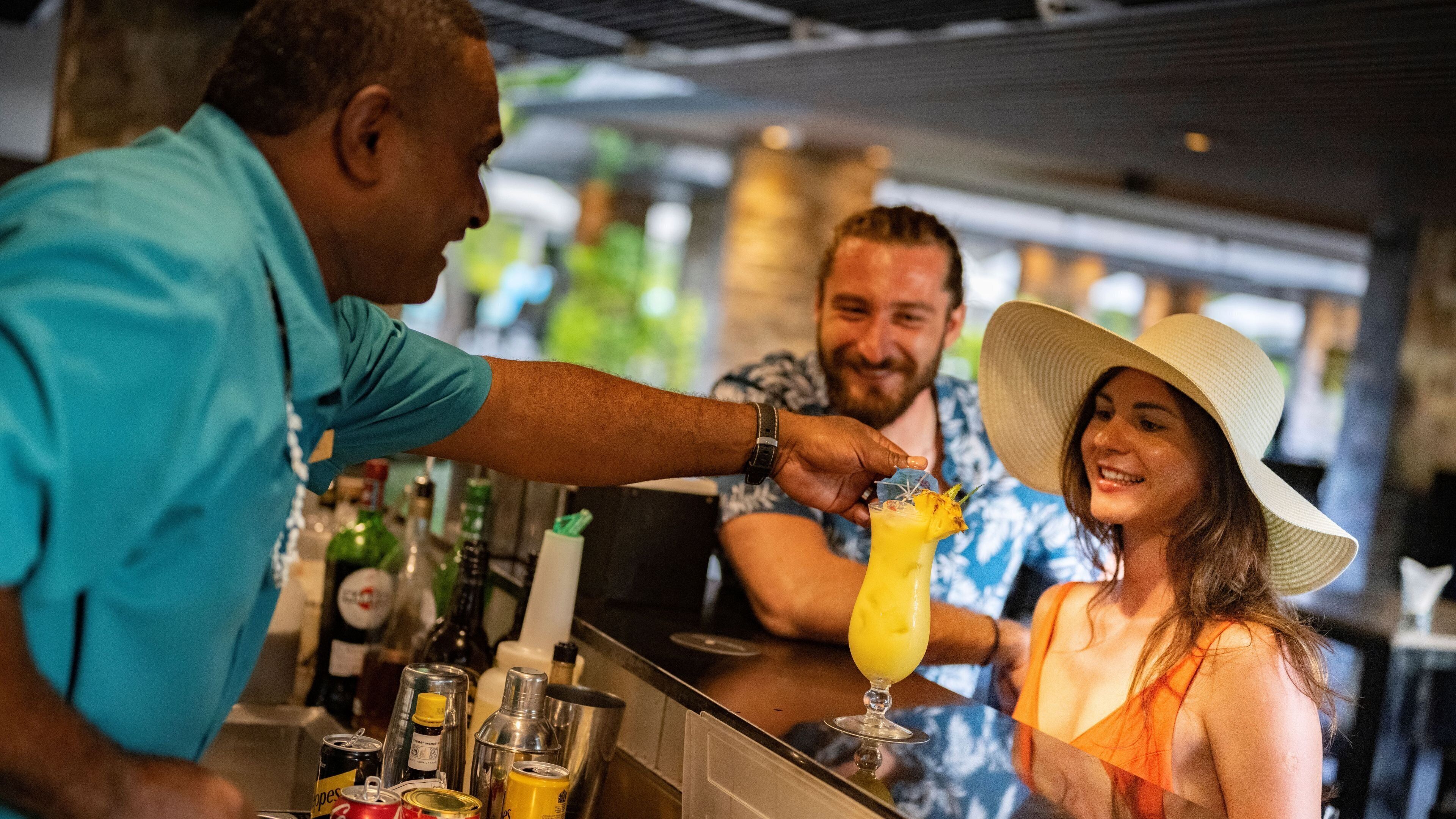Un bar con un cantinero preparando una bebida, una mujer con sombrero de paja y un hombre con camisa azul.