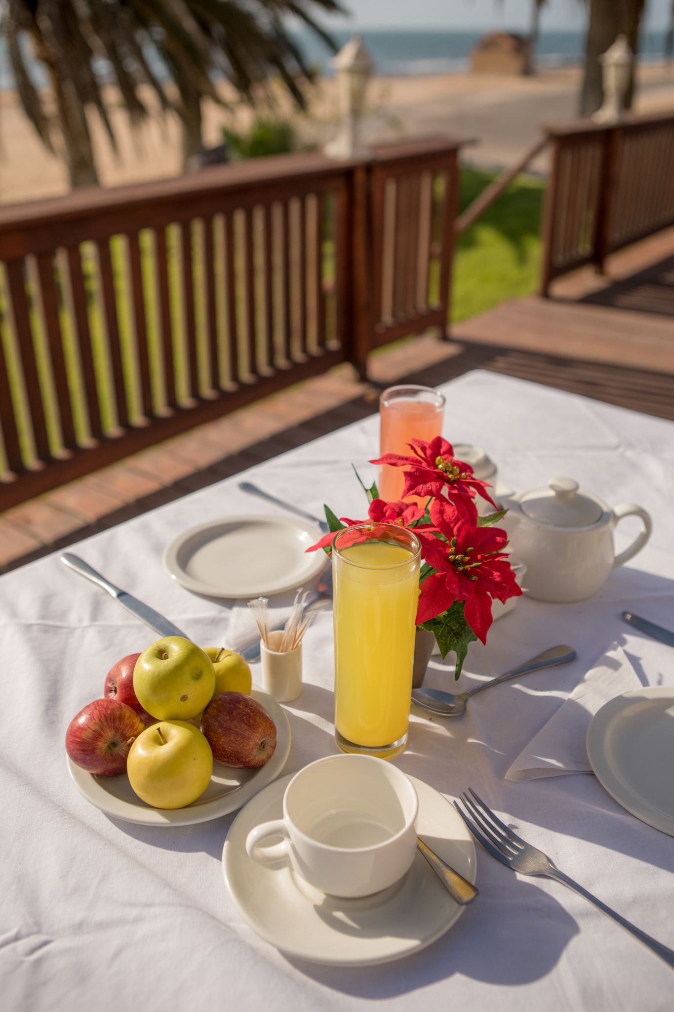 A table set with breakfast items including fruit, a drink, and a teapot, with a view of the ocean in the background.