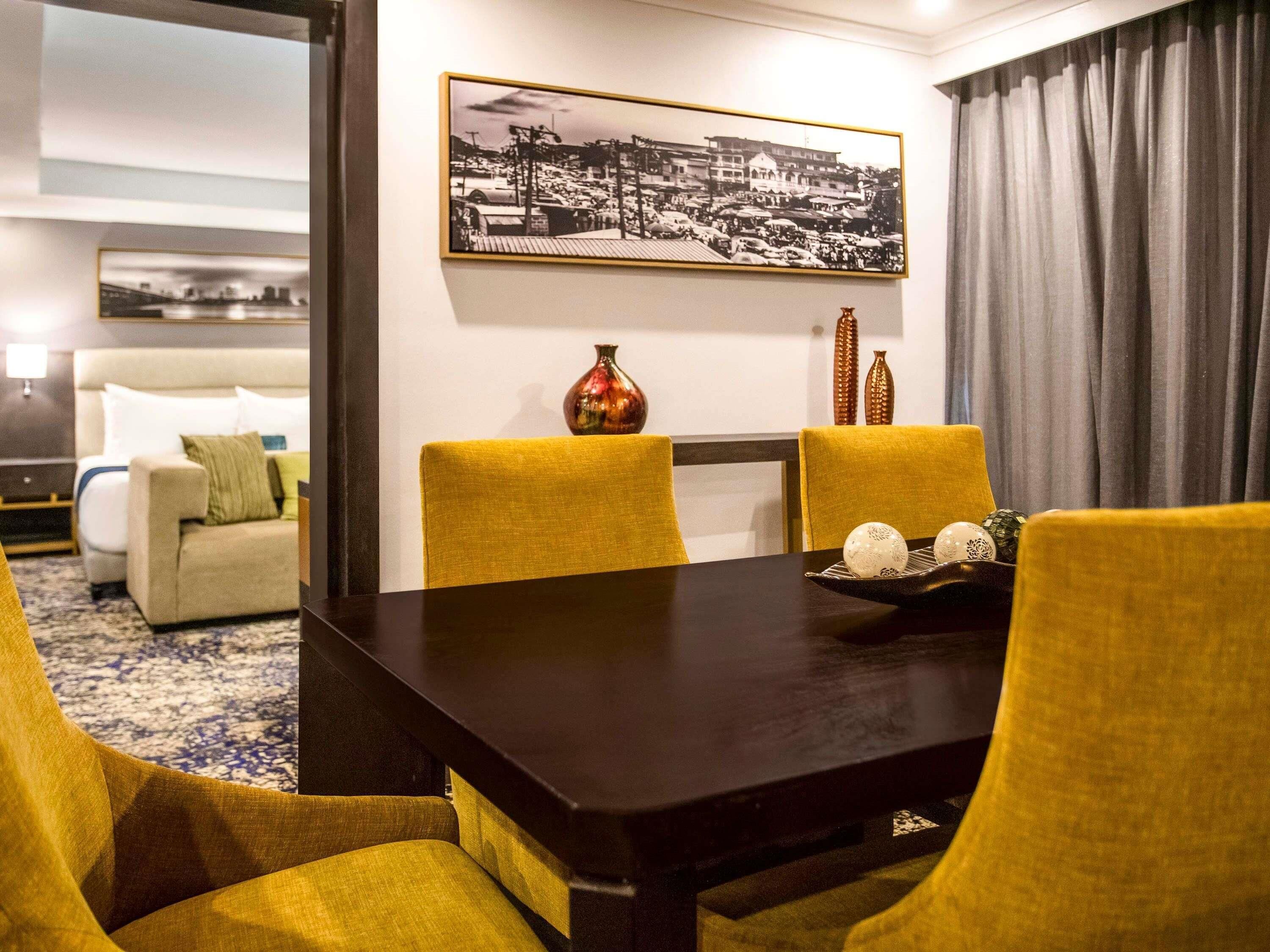 Hotelkamer met een eettafel, gele stoelen, een bank, een bed en een lijst met een stadsgezicht aan de muur.