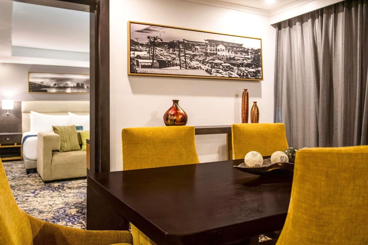 A hotel room with a dining table, yellow chairs, a sofa, a bed, and a framed cityscape picture on the wall.