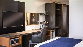 Habitación de hotel con cama, escritorio, silla, televisor, espejo y minibar.