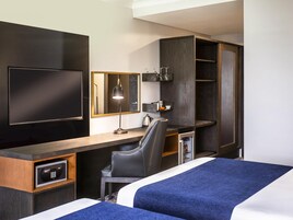 Habitación de hotel con cama, escritorio, silla, televisor, espejo y minibar.