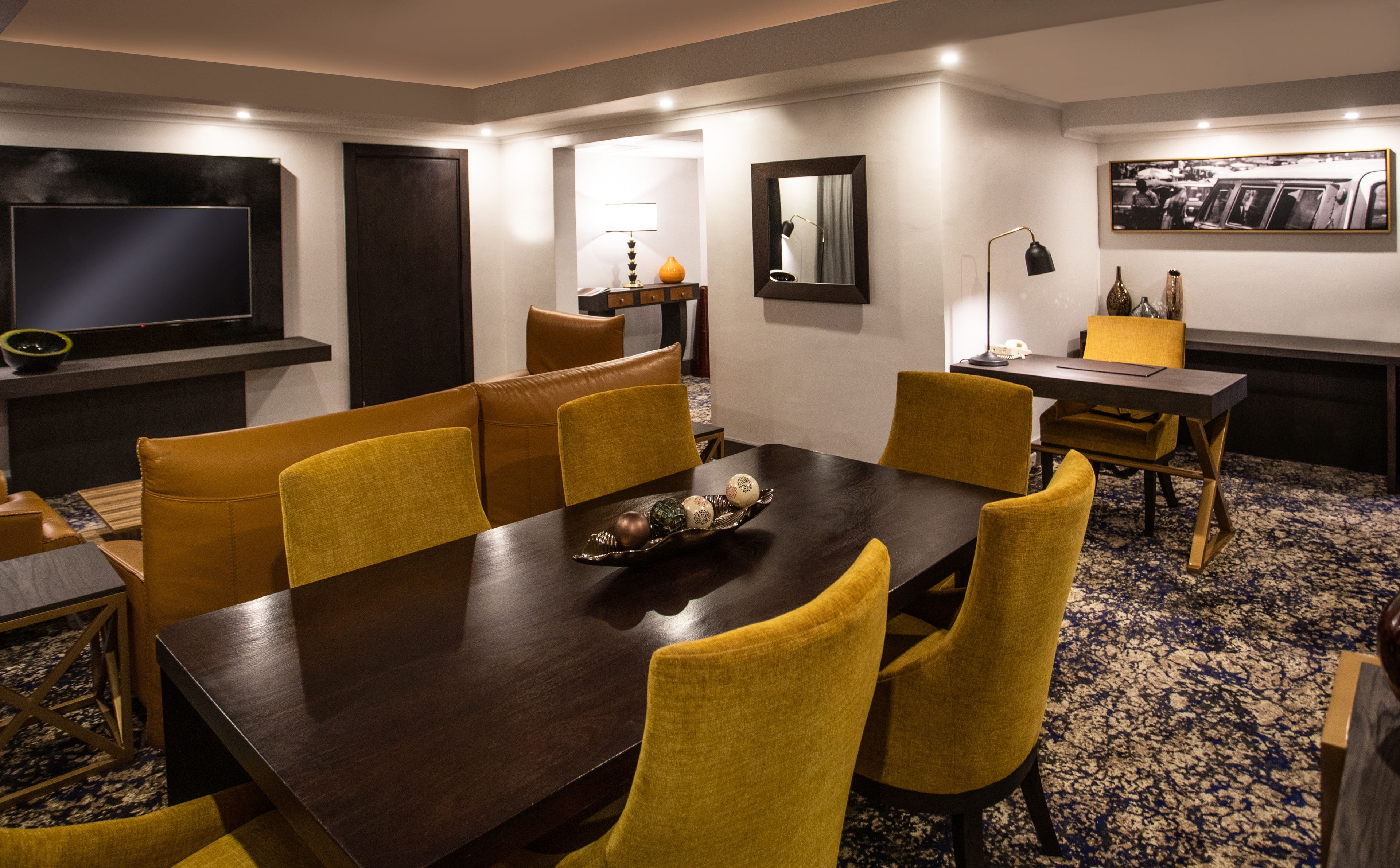 A conference room with a dark wooden table, yellow chairs, a flat-screen TV, a mirror, and a framed photo on the wall.