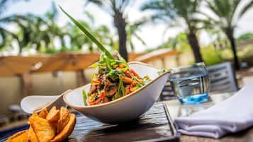 A bowl of food with a spoon, a glass of water, and a napkin on a table with a view of palm trees.