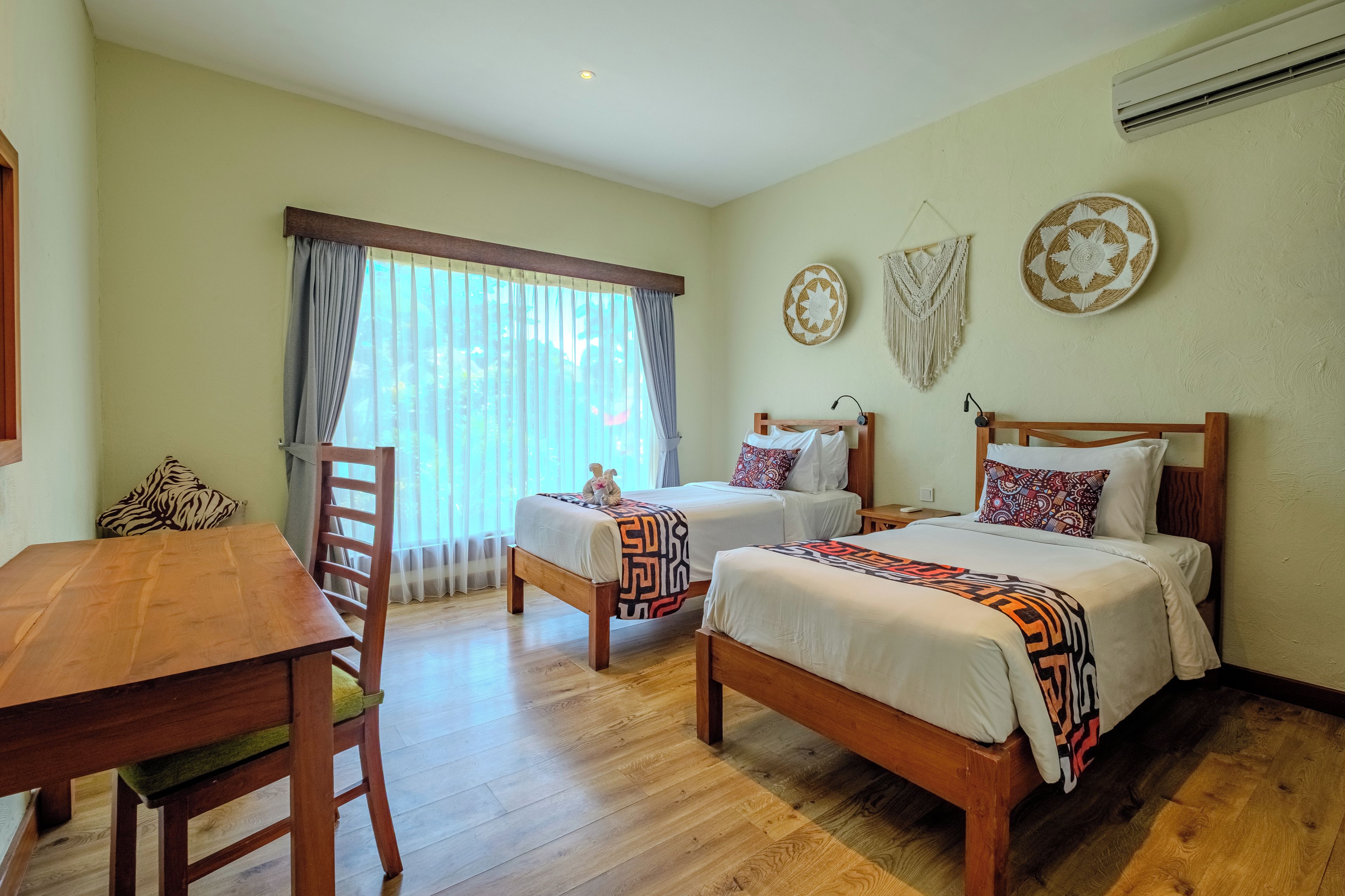 A hotel room with two beds, a wooden table, a chair, and a large window with curtains.