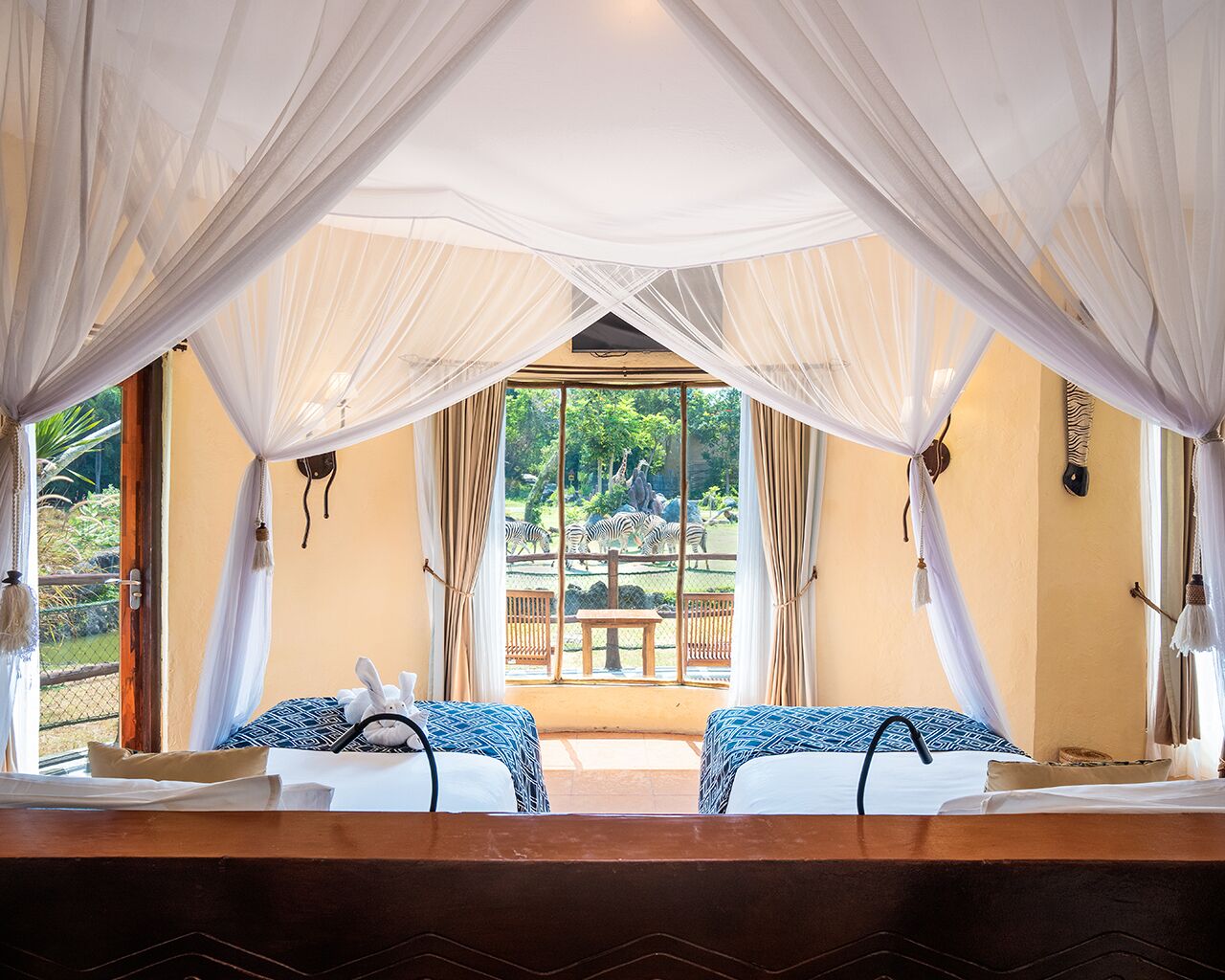 A room with two beds, a view of zebras outside, and a canopy over the beds.