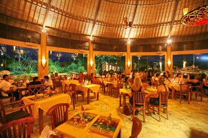 A restaurant with multiple tables and chairs, a thatched roof, and large windows overlooking a garden.