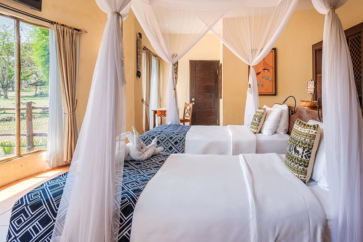A room with two beds, white bedding, and a canopy bed. There is a window with curtains, a chair, and a painting on the wall.