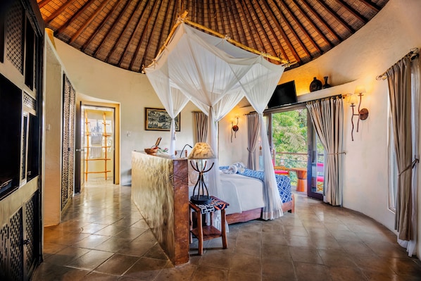 A room with a bed, a nightstand, a lamp, a chair, a TV, and a window with curtains. - Mara River Safari Lodge (Gianyar)
