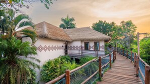 A wooden bridge leading to a thatched-roof building with patterned walls, surrounded by lush greenery.