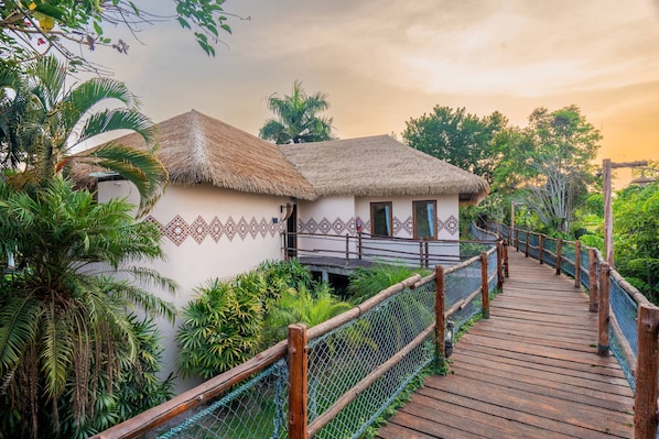 A wooden bridge leading to a thatched-roof building with patterned walls, surrounded by lush greenery.