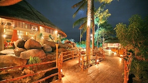 A wooden deck with a thatched-roof building, palm trees, and a wooden fence.