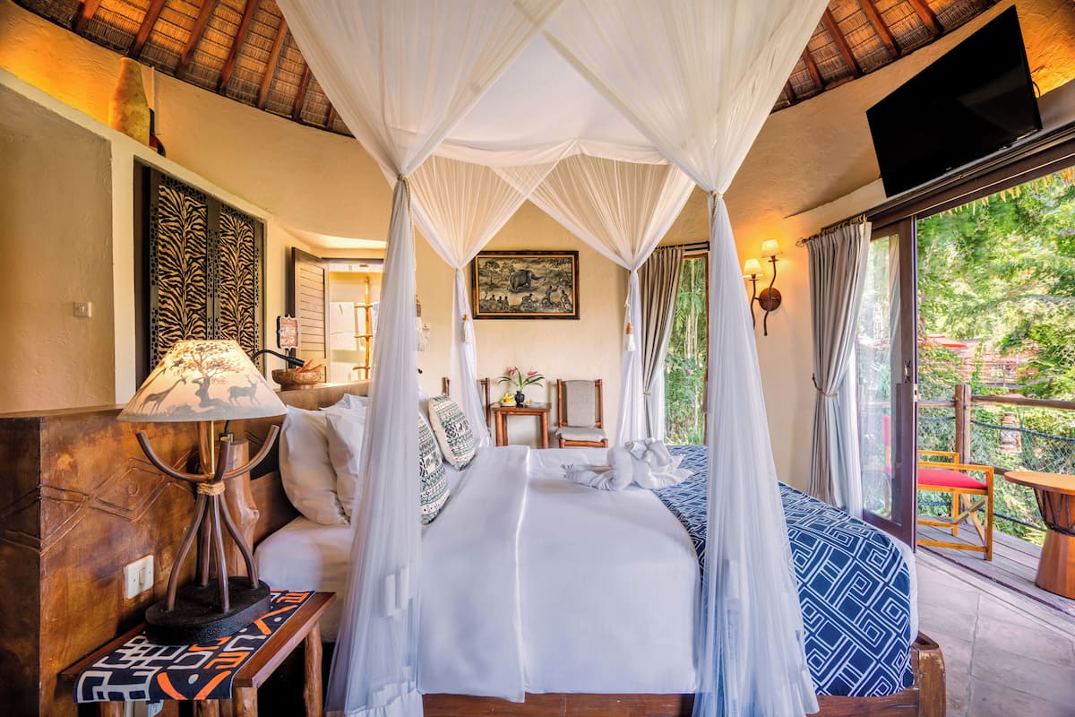 A bedroom with a canopy bed, a bedside table with a lamp, a chair, and a view of the outdoors.