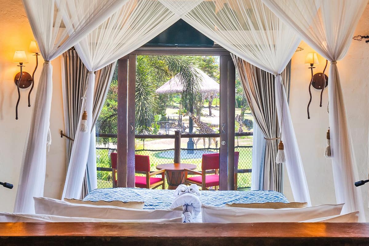 A room with a bed, a view of an outdoor area with a giraffe, and a table with chairs.