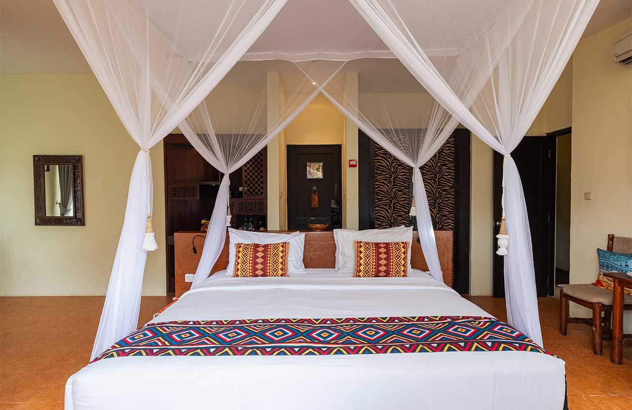 A bed with a canopy, pillows, and a patterned bedspread in a room with a wooden floor and a door in the background.