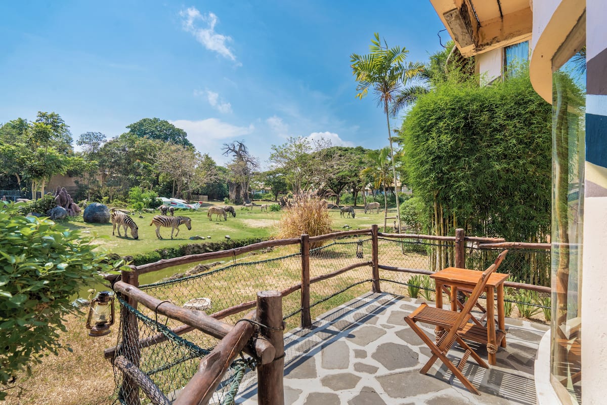 A patio with a wooden table and chairs, a stone floor, and a view of a zoo with zebras and giraffes.