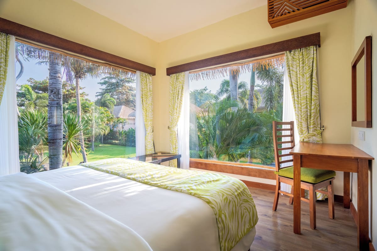A bedroom with a large bed, a wooden desk, and a view of a tropical garden.