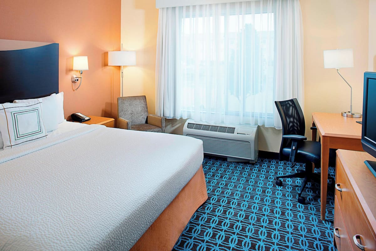 A hotel room with a large bed, a desk with a computer, a chair, a desk lamp, and a window with curtains.