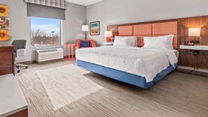 A modern hotel room with a large bed, a desk, a chair, a radiator, a window with a view, and a painting on the wall. - Hampton Inn Denver Northeast-Brighton (Brighton)