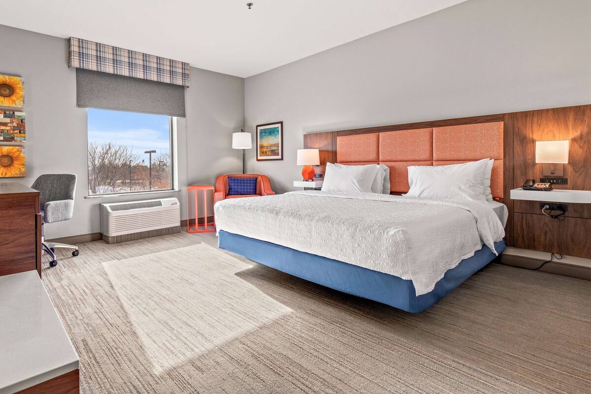 A modern hotel room with a large bed, a desk, a chair, a radiator, a window with a view, and a painting on the wall.