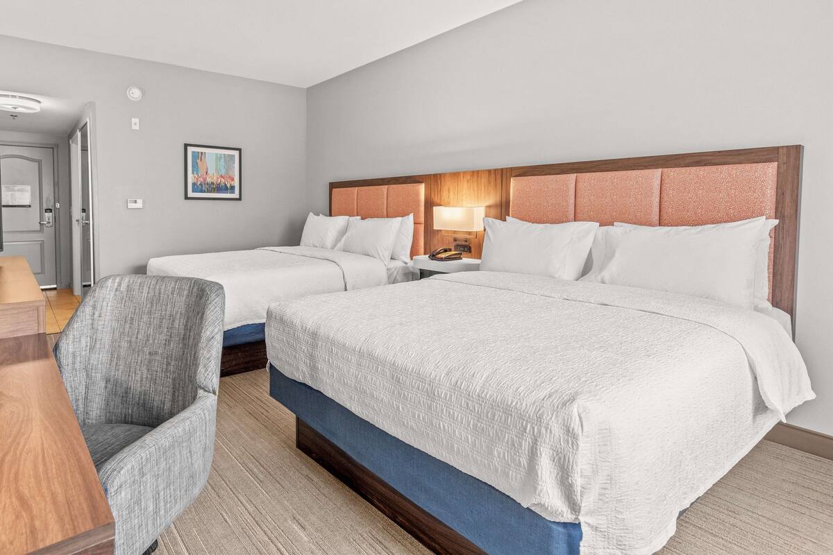 A hotel room with two beds, a desk, a chair, and a nightstand with a lamp.