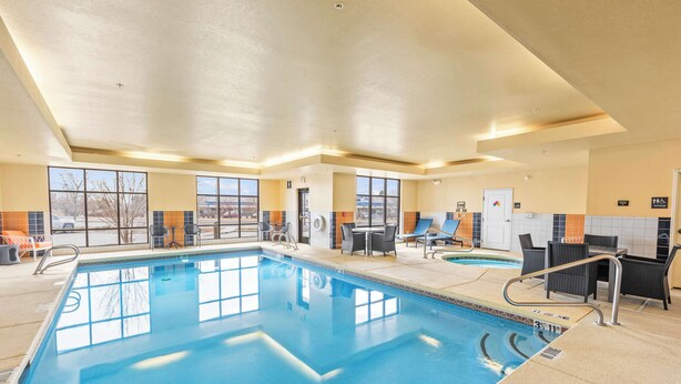 An indoor swimming pool with clear water, surrounded by lounge chairs and tables, large windows, and a tiled wall. - Hampton Inn Denver Northeast-Brighton (Brighton)