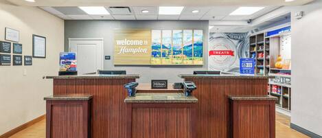 Zona de recepción de hotel con un cartel que dice 'Welcome to Hampton', una sección de 'Treats' y un cartel de 'Hilton Honors'.