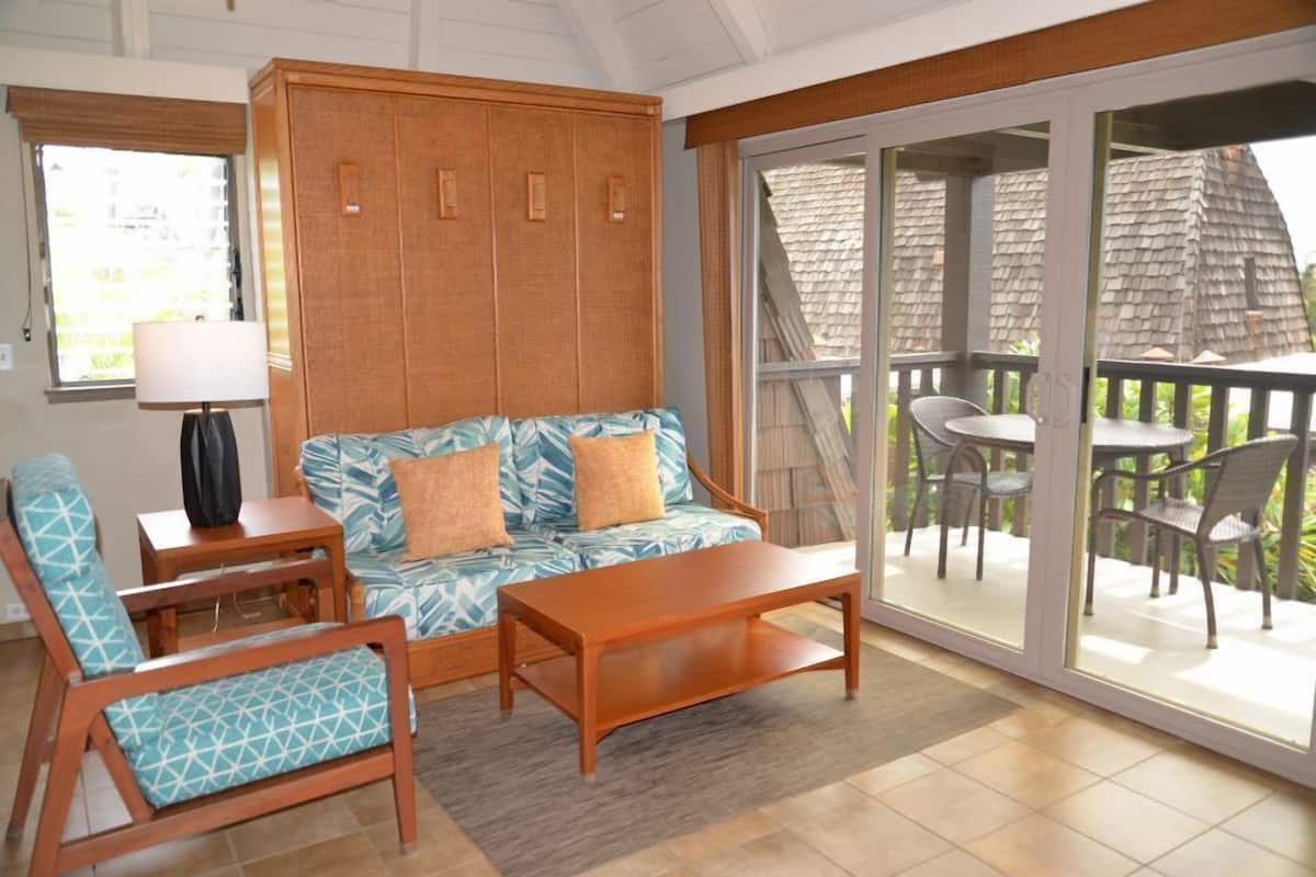 A living room with a sofa, chairs, a coffee table, a lamp, and a sliding glass door leading to a balcony with outdoor furniture.