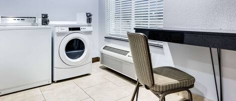 A laundry room with a front-loading washer and dryer, a chair, and a window with blinds.