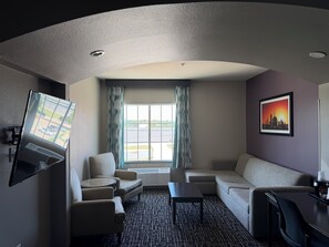 Ein Hotelzimmer mit gewölbter Decke, einem an der Wand montierten Flachbildfernseher, einer Couch, zwei Sesseln, einem Couchtisch und Blick auf den Parkplatz durch ein mit Vorhängen versehenes Fenster.