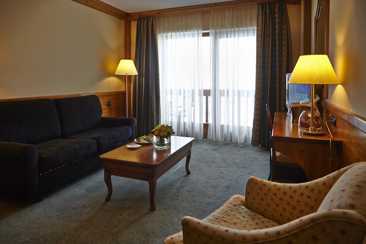 A hotel room with a dark sofa, a wooden coffee table with a vase of flowers, a patterned armchair, and a wooden desk with a lamp.