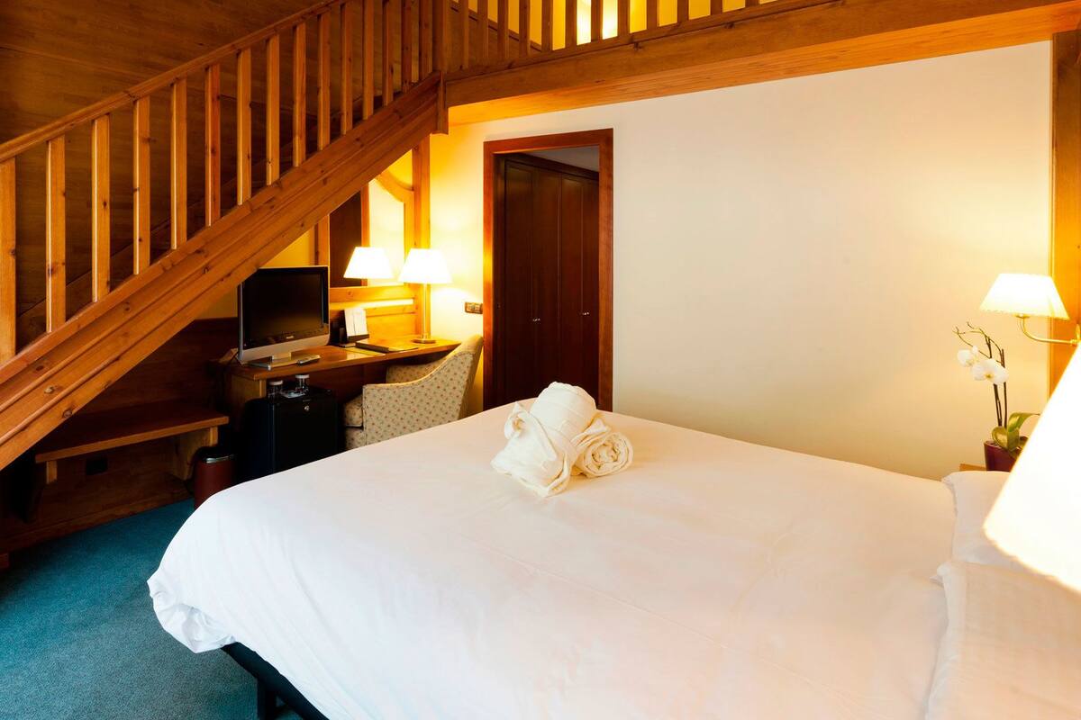 A hotel room with a wooden staircase, a desk with a computer, a bed with a rolled towel, and two bedside lamps.