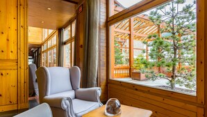 A cozy cabin interior with wooden walls and large windows, a comfortable seating area, and a view of a wooden deck and pine tree outside. - Sport Village (Soldeu)
