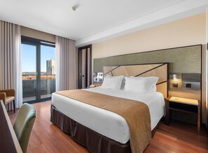 A hotel room with a large bed, a desk, a chair, a nightstand, and a view of the city. - Axis Porto Business & Spa Hotel (Matosinhos)