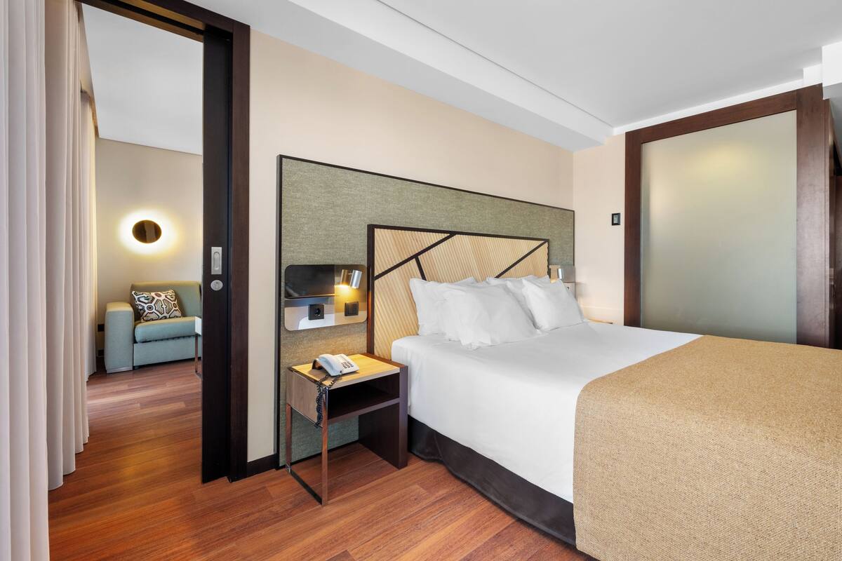 A hotel room with a bed, bedside table, lamp, and a view of another room.