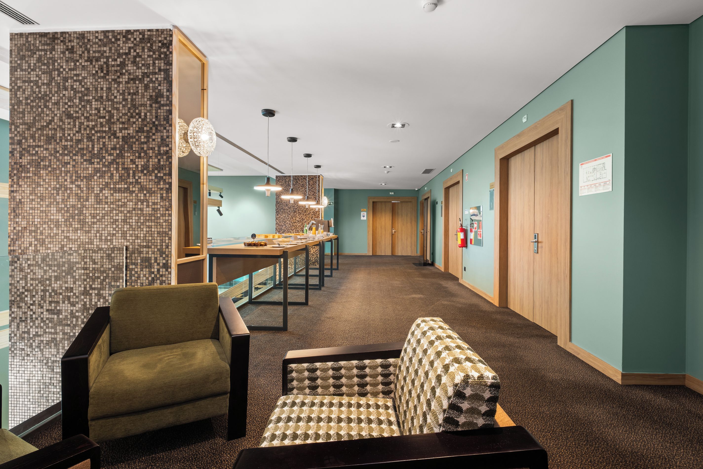 A hotel corridor with a seating area, a long table with chairs, and a patterned bench.