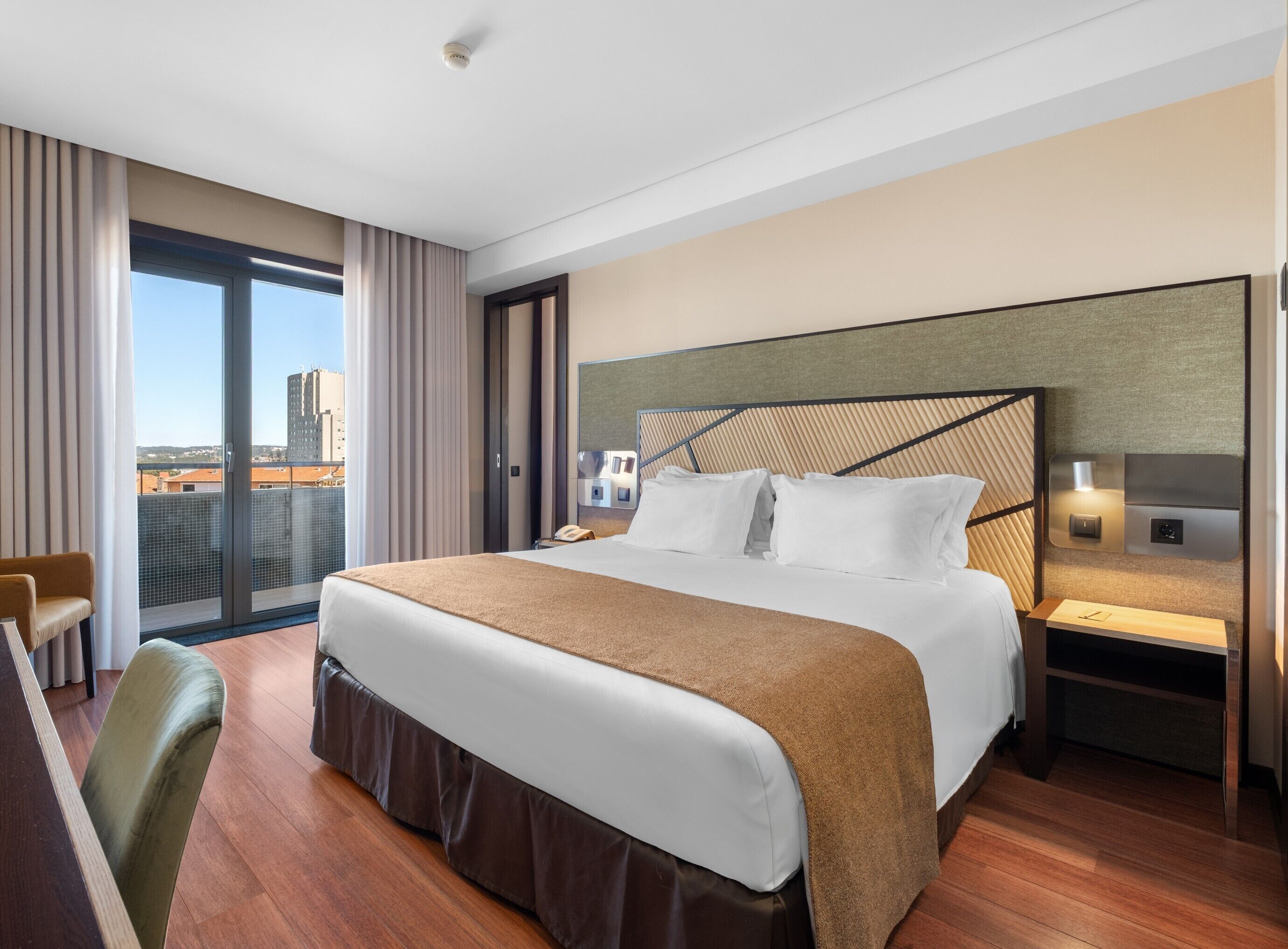A hotel room with a large bed, a desk, a chair, a nightstand, and a view of the city.