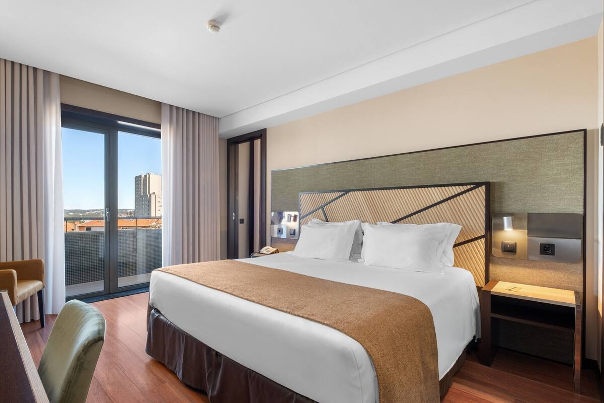 A hotel room with a large bed, a desk, a chair, a nightstand, and a view of the city.