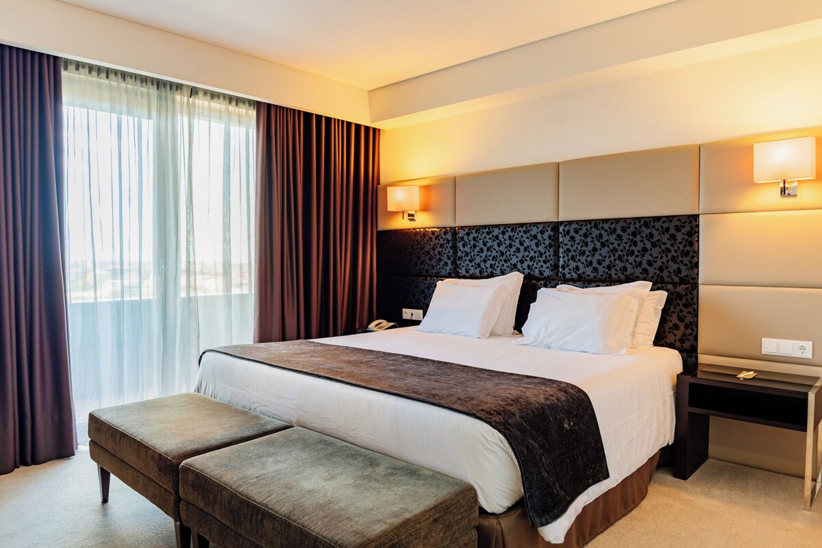 A hotel room with a large bed, two bedside tables, a bench, and a window with curtains.