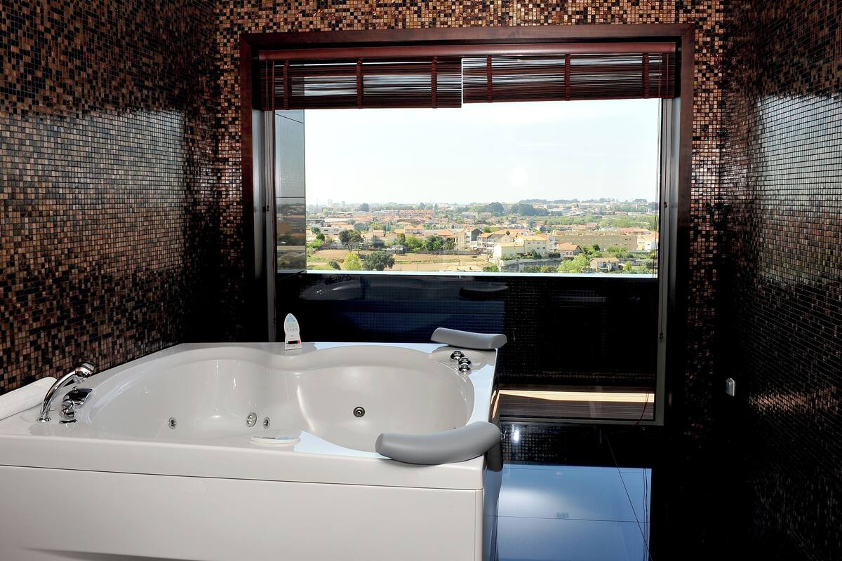 A bathroom with a large bathtub, a window with a view of a cityscape, and mosaic tile walls.