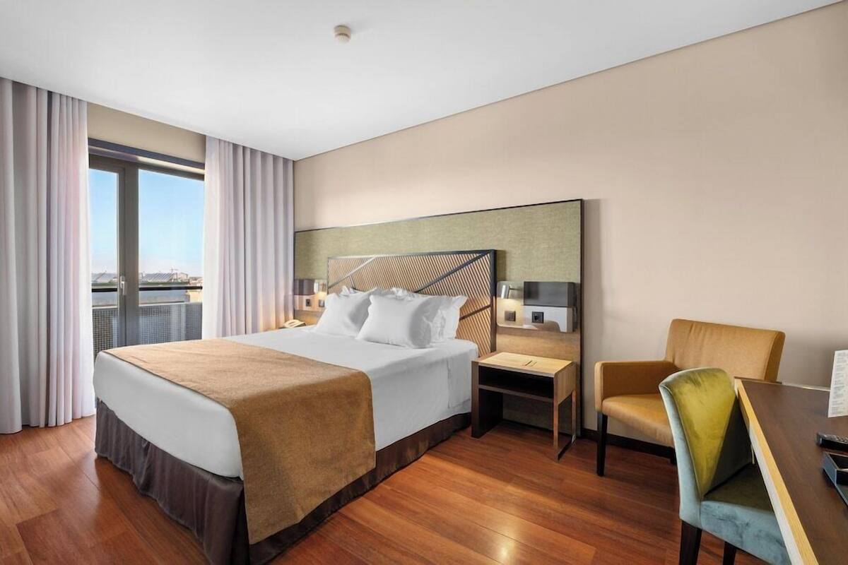 A modern hotel room with a large bed, a desk, a chair, and a view of the cityscape.