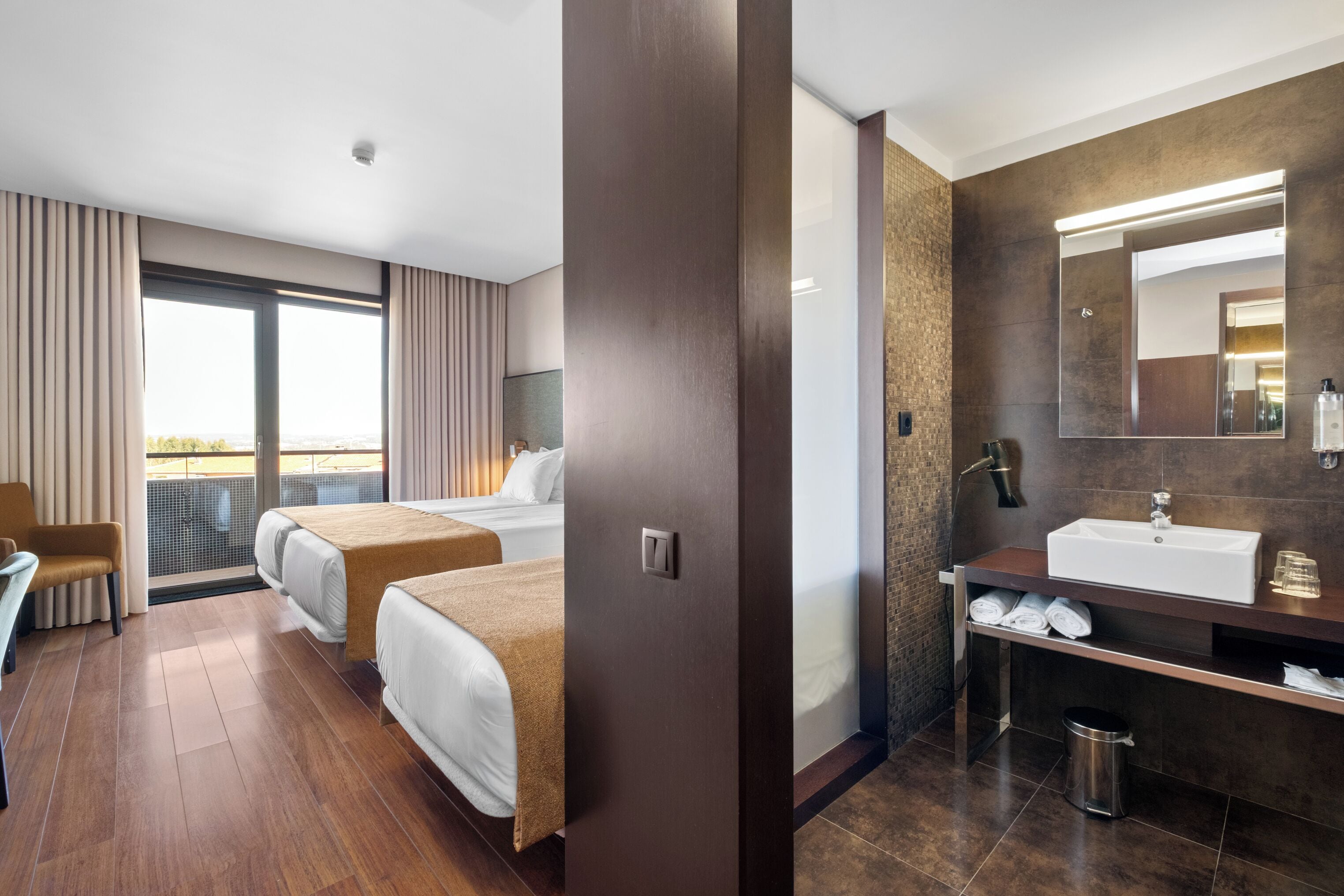 A hotel room with two beds, a sitting area, a bathroom with a large mirror, and a view of the outdoors.