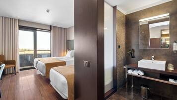 Quarto de hotel com duas camas, um espaço de estar, banheiro com um espelho grande e vista para o exterior.
